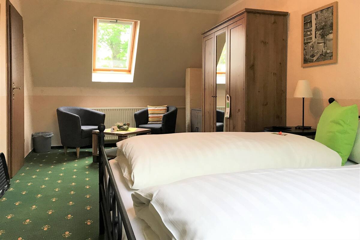 Habitación de hotel con dos camas, un escritorio, dos sillas, una ventana, un radiador, una lámpara y un cuadro en la pared.