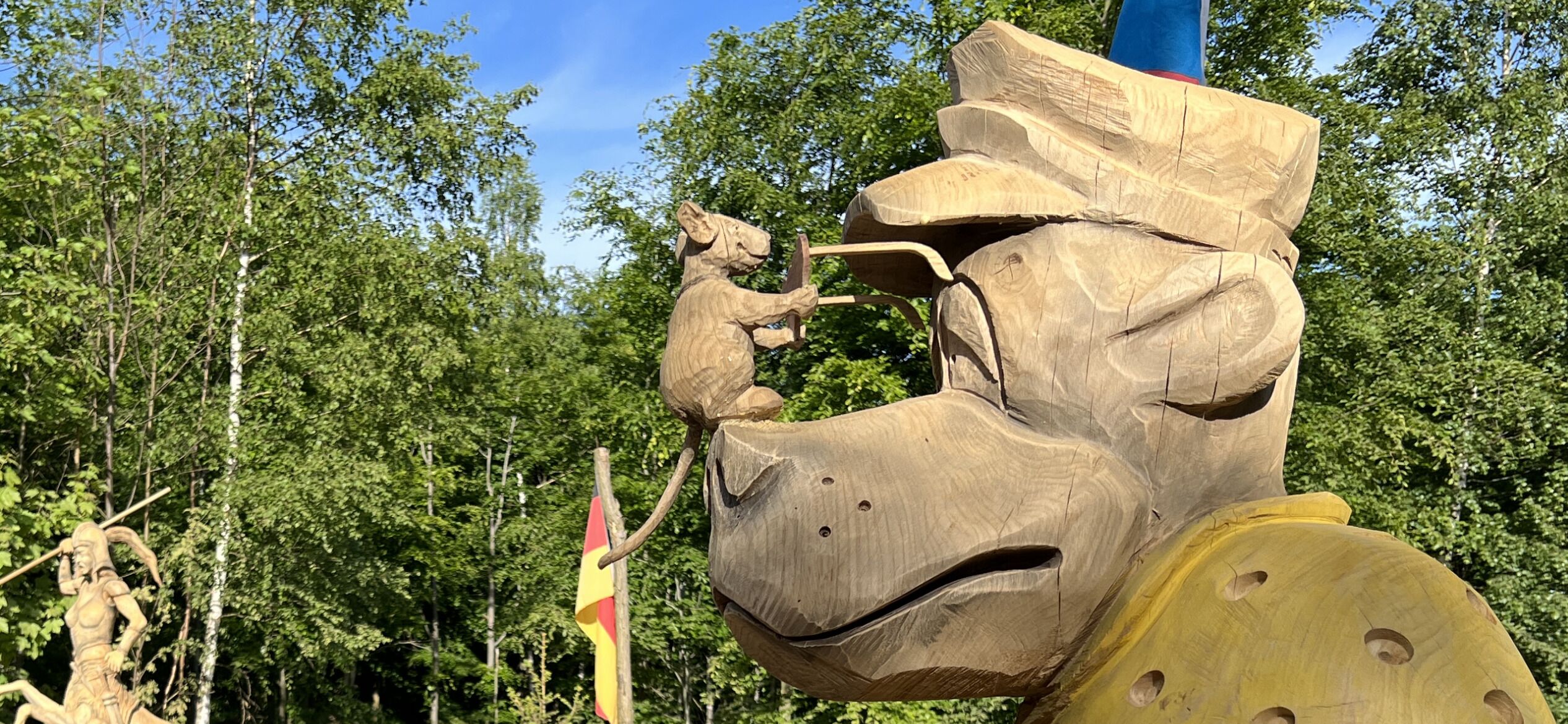 A wooden sculpture of a horse's head with a rider, set against a backdrop of trees and a flag.
