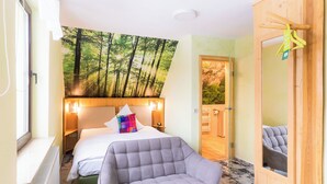 Un dormitorio con una cama, un sillón gris, un gran mural de un bosque en la pared y una puerta de madera.