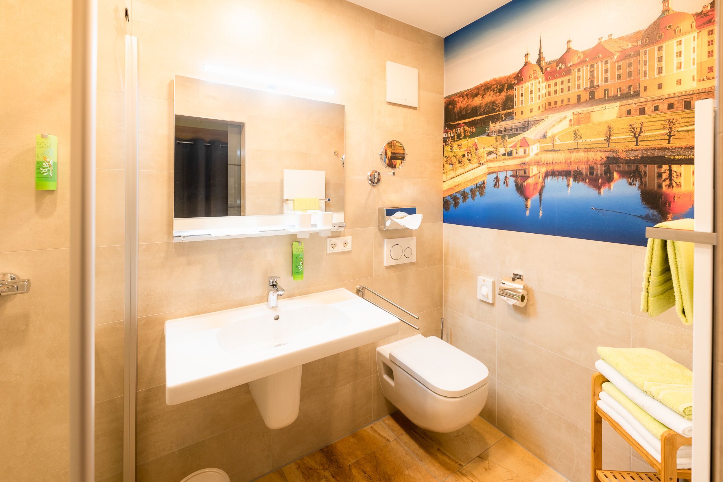 A bathroom with a sink, toilet, and a large mirror. There is a towel rack with towels and a painting of a building on the wall.