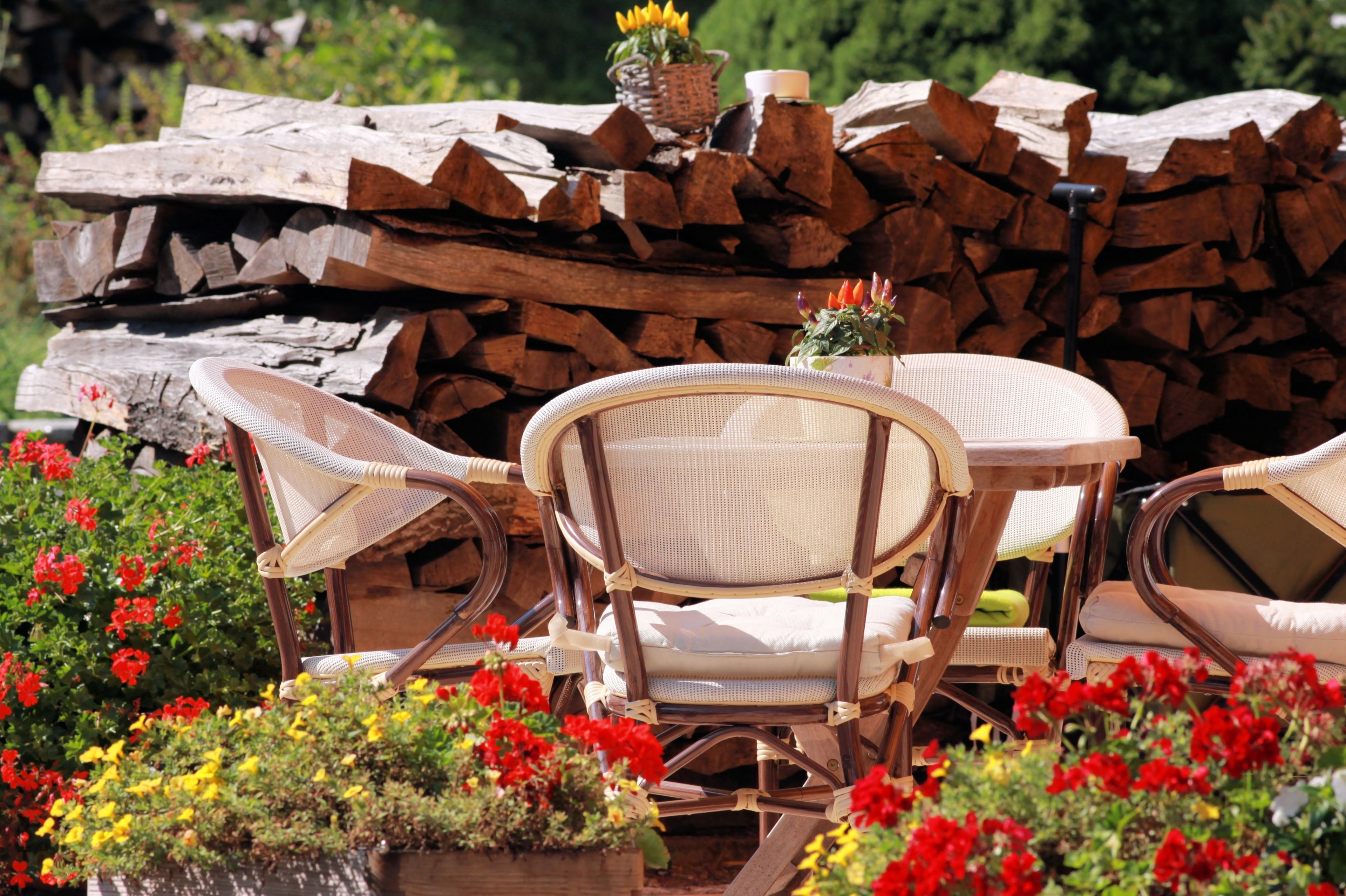Ein rustikaler Sitzbereich im Freien mit Korbstühlen und einem Tisch, umgeben von bunten Blumen und einem Holzstoß.