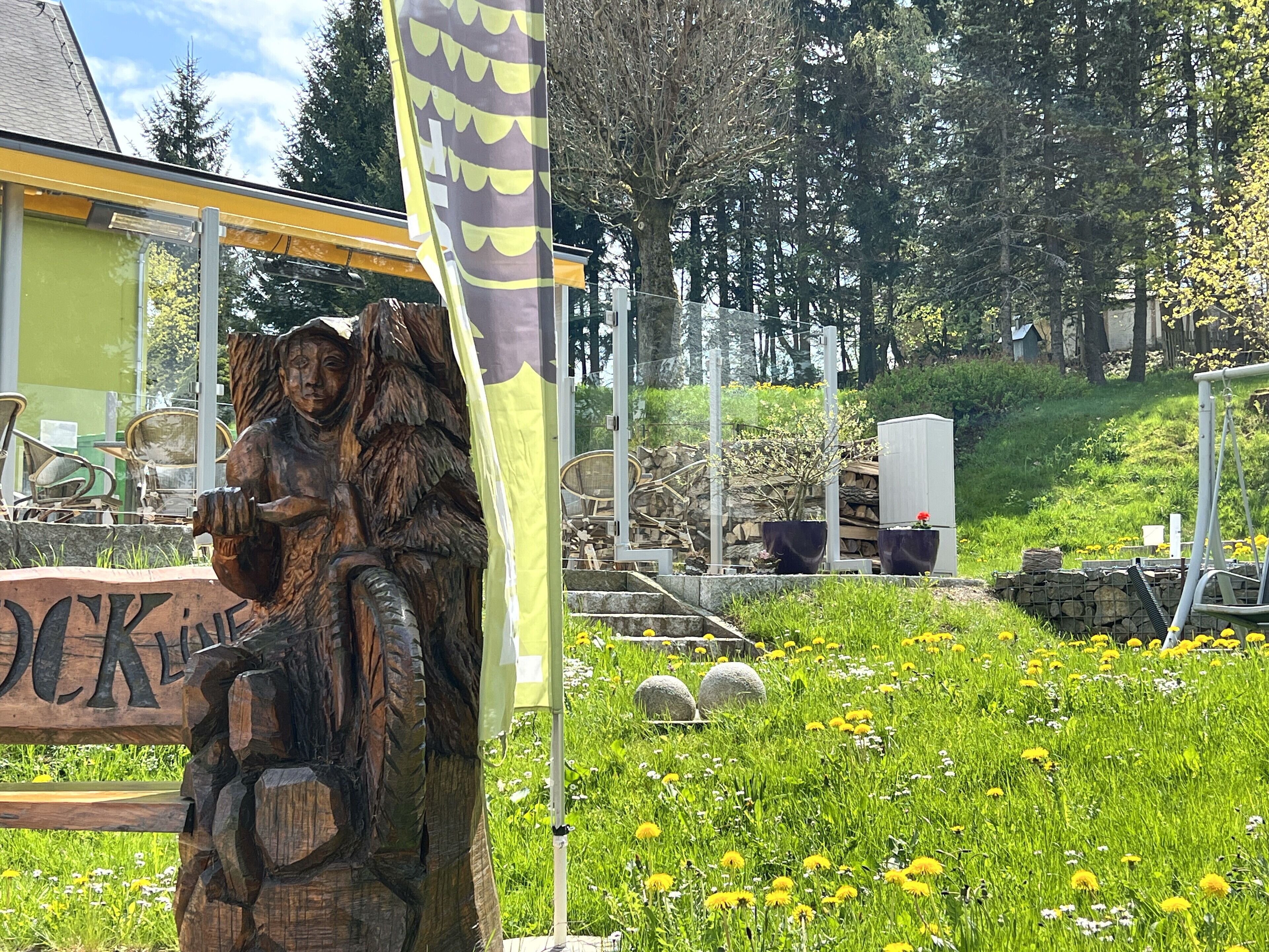 A wooden sculpture of a figure holding a staff, a yellow flag, and a sign with the word "LOCK" in a garden setting.
