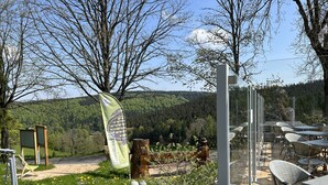 Un espacio al aire libre con mesas y sillas, rodeado de césped con flores amarillas y vistas a una colina boscosa.