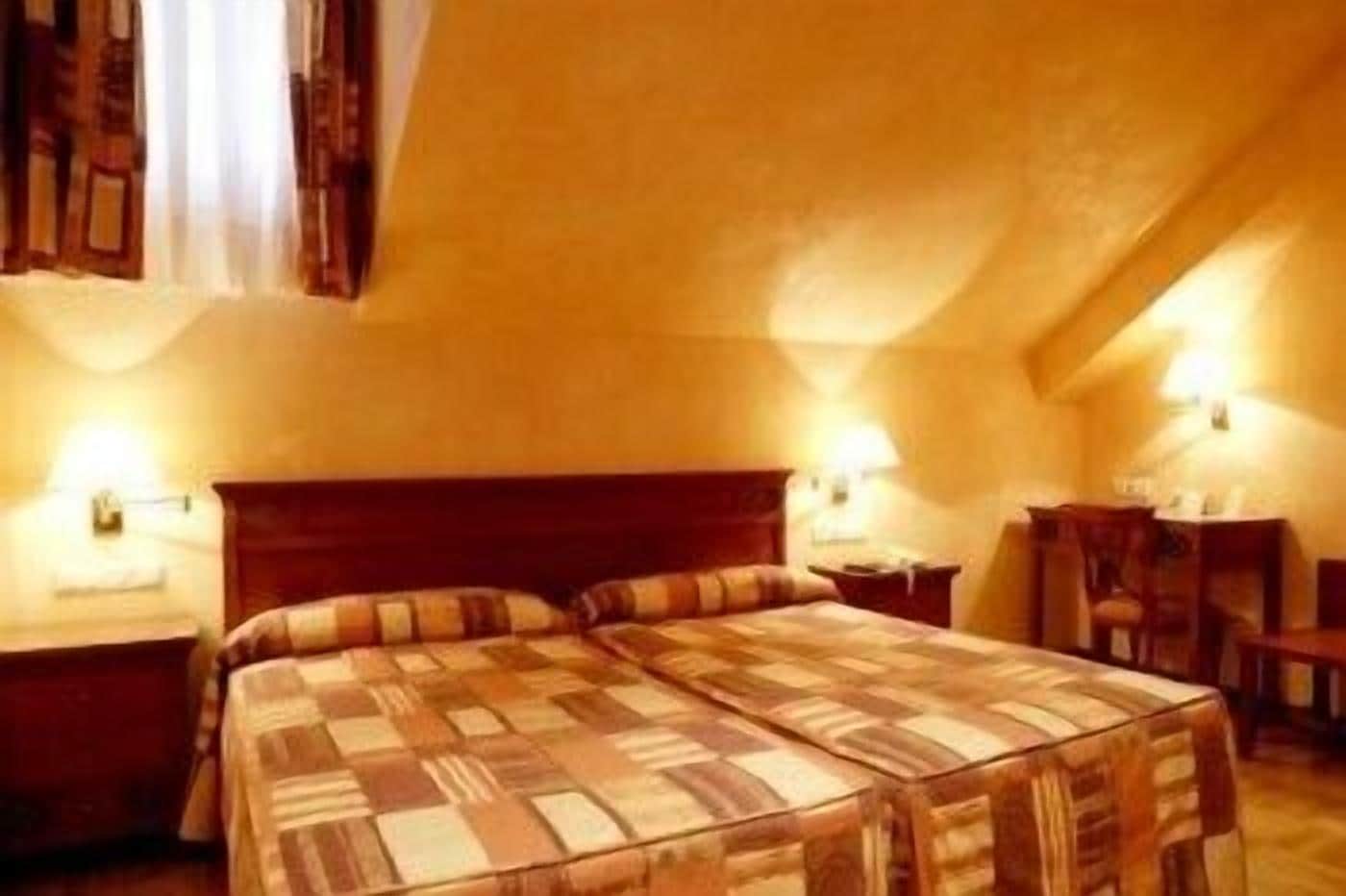 A cozy, single-room hotel bedroom with a large bed, a nightstand, a bedside table, and a chair.