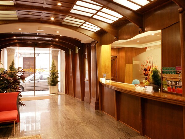 A hotel lobby with a reception desk, a red seating area, and a large window with a view of the street.