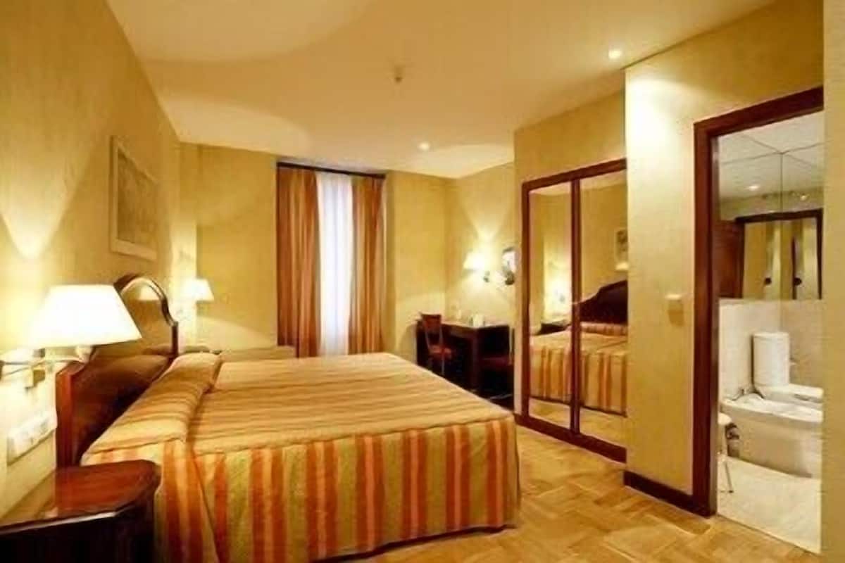 A hotel room with two beds, a desk, a chair, a lamp, a mirror, and a bathroom visible through an open door.