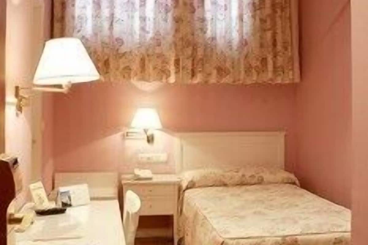 A bedroom with a single bed, a bedside table, a lamp, and a window with curtains.