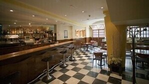 A bar area with stools and a checkered floor, a seating area with tables and chairs, and a view of the bar through a window. - Cordón (Burgos)