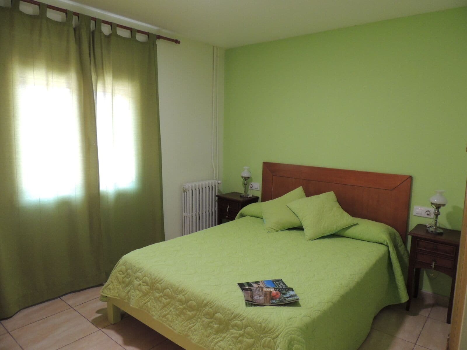Habitació amb llit, parets verdes, radiador i finestra amb cortines.
