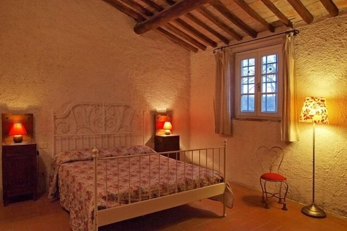 Un dormitorio con una cama, dos mesitas de noche con lámparas, una silla y una ventana con cortinas.
