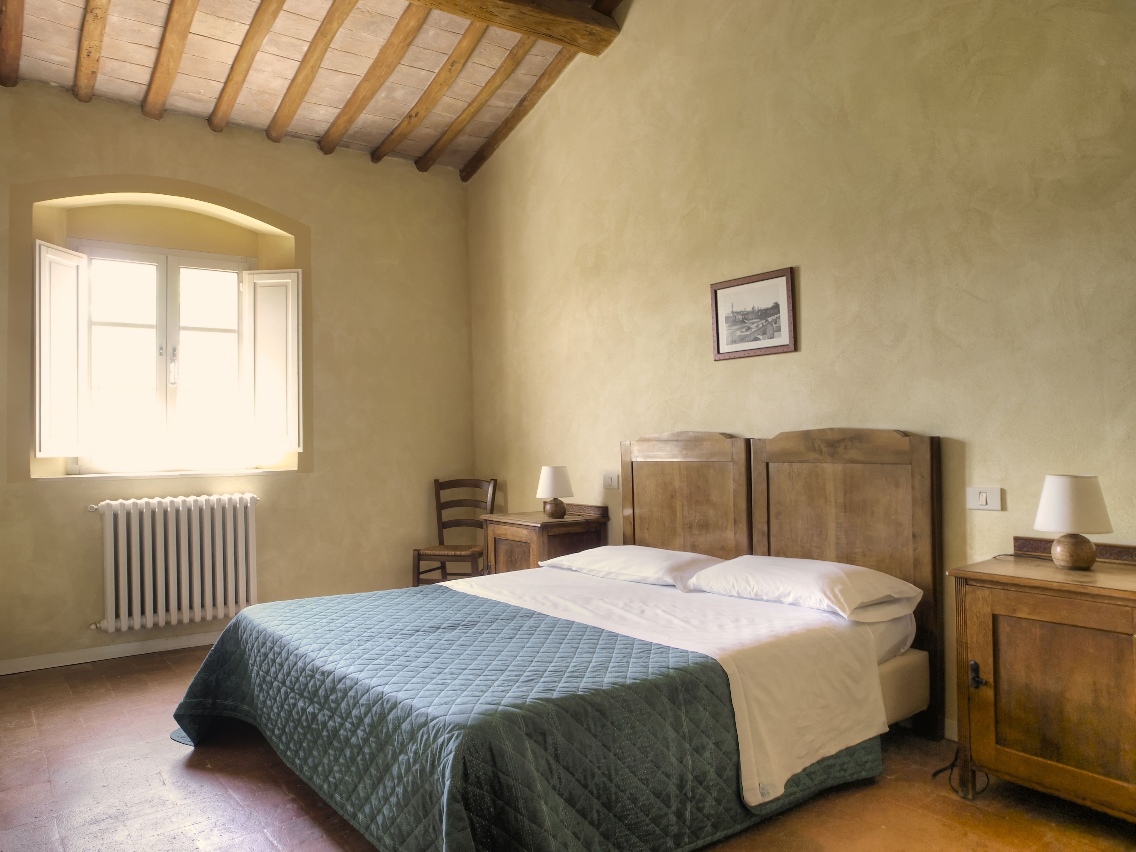 A bedroom with a bed, a chair, a nightstand, a lamp, a radiator, and a window.