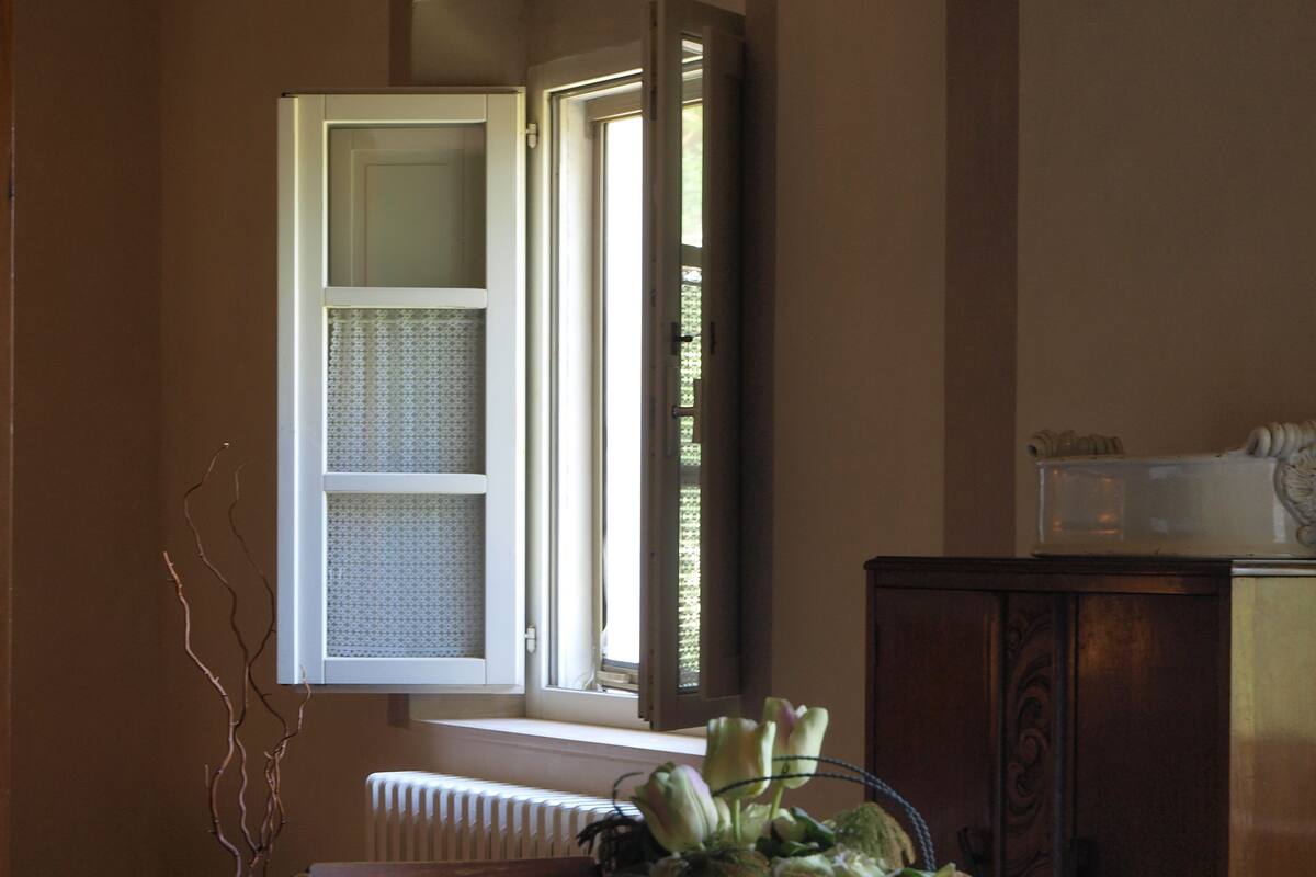 Un área de comedor con una mesa, sillas, un jarrón de flores, un radiador y una ventana con contraventanas abiertas.