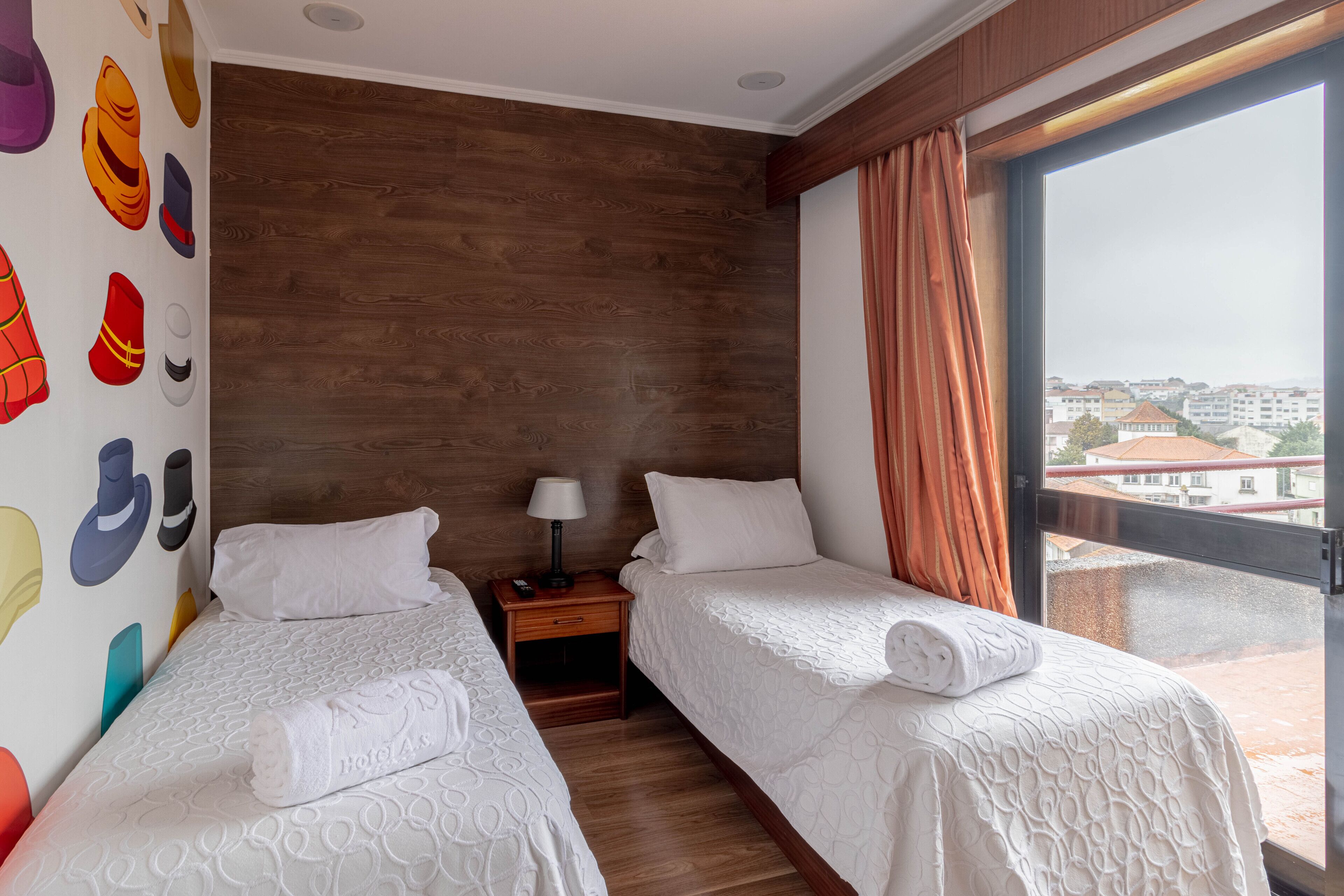 A hotel room with two beds, a wooden wall, a nightstand with a lamp, and a large window with curtains.