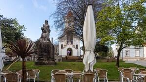An outdoor cafe area with wicker chairs and tables, a statue, and a building with a tower in the background.