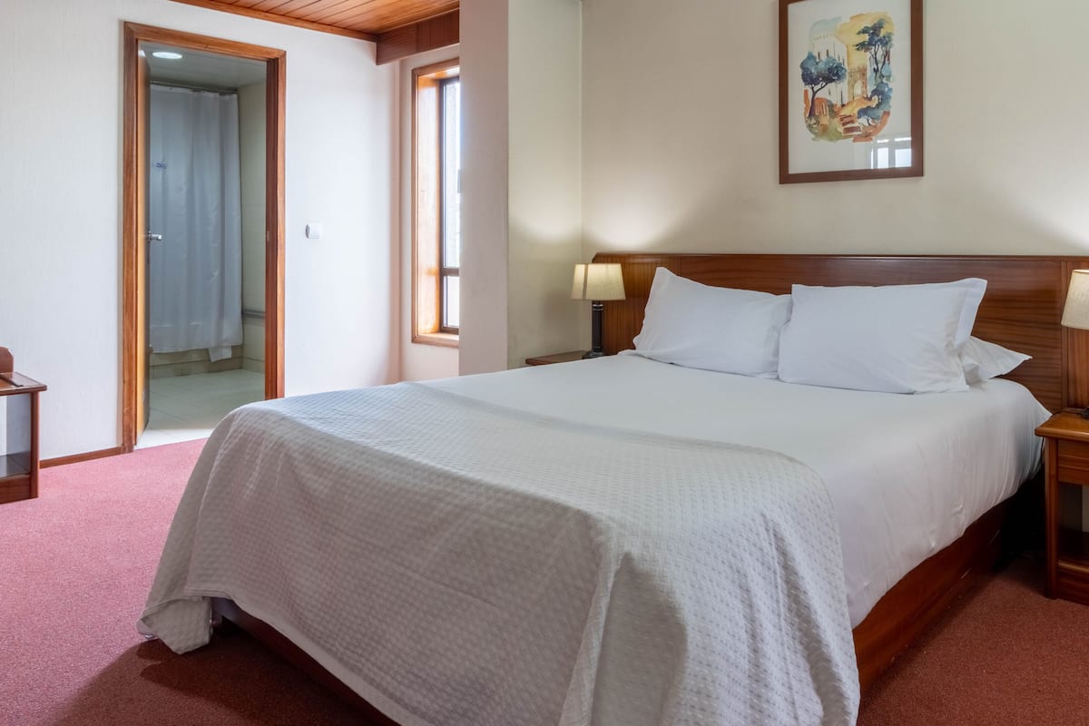 A hotel room with a large bed, a wooden headboard, a nightstand with a lamp, and a painting on the wall.