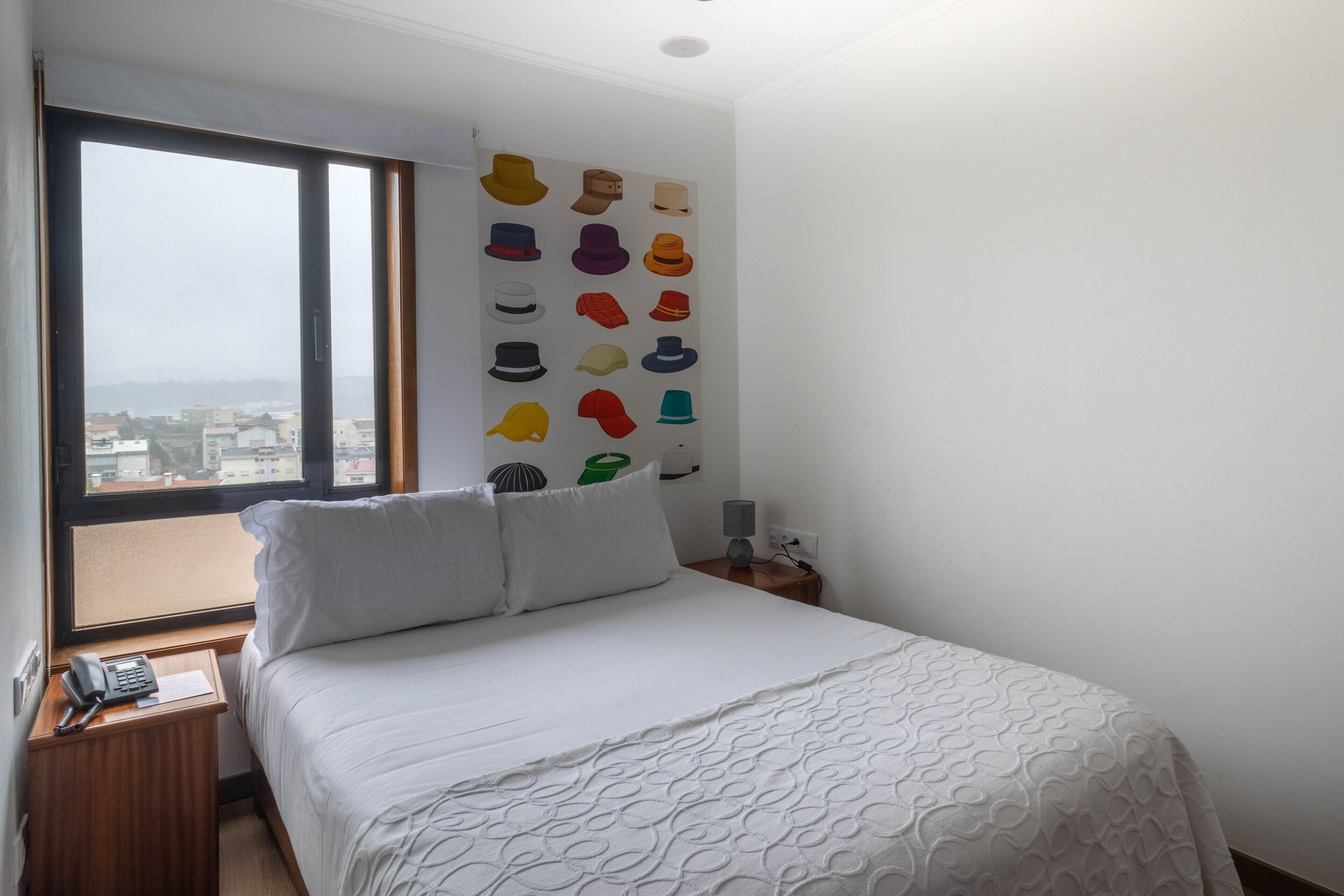 A hotel room with a large window, a bed with a patterned bedspread, a wall-mounted hat display, and a bedside table with a lamp and phone.