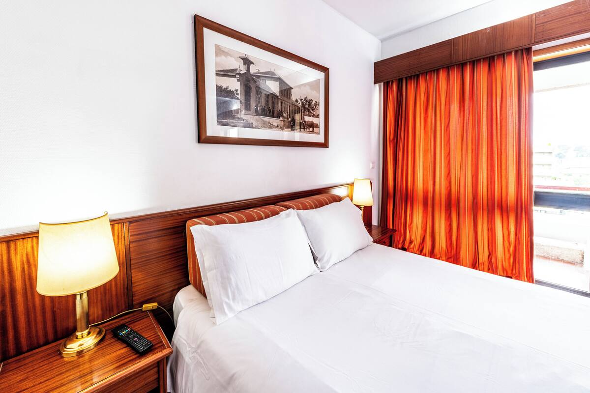 A hotel room with a bed, two bedside lamps, a nightstand, a framed picture on the wall, and a window with curtains.