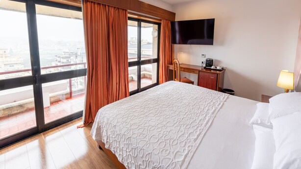 A hotel room with a large bed, a desk, a chair, a television, and a view of the city through a large window. - AS São João da Madeira (Sao Joao da Madeira)