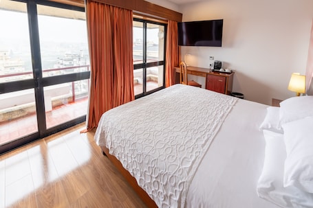 A hotel room with a large bed, a desk, a chair, a television, and a view of the city through a large window.