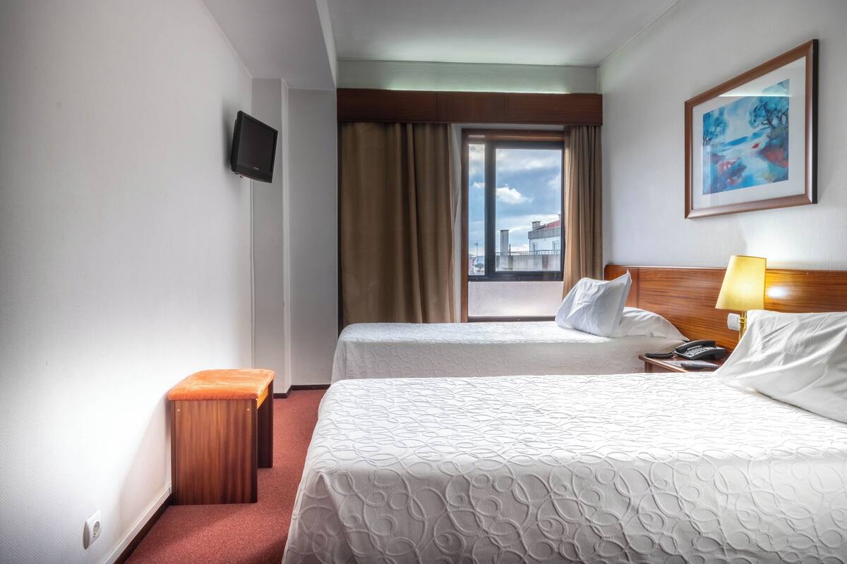 A hotel room with two beds, a TV, a painting, and a window with curtains.