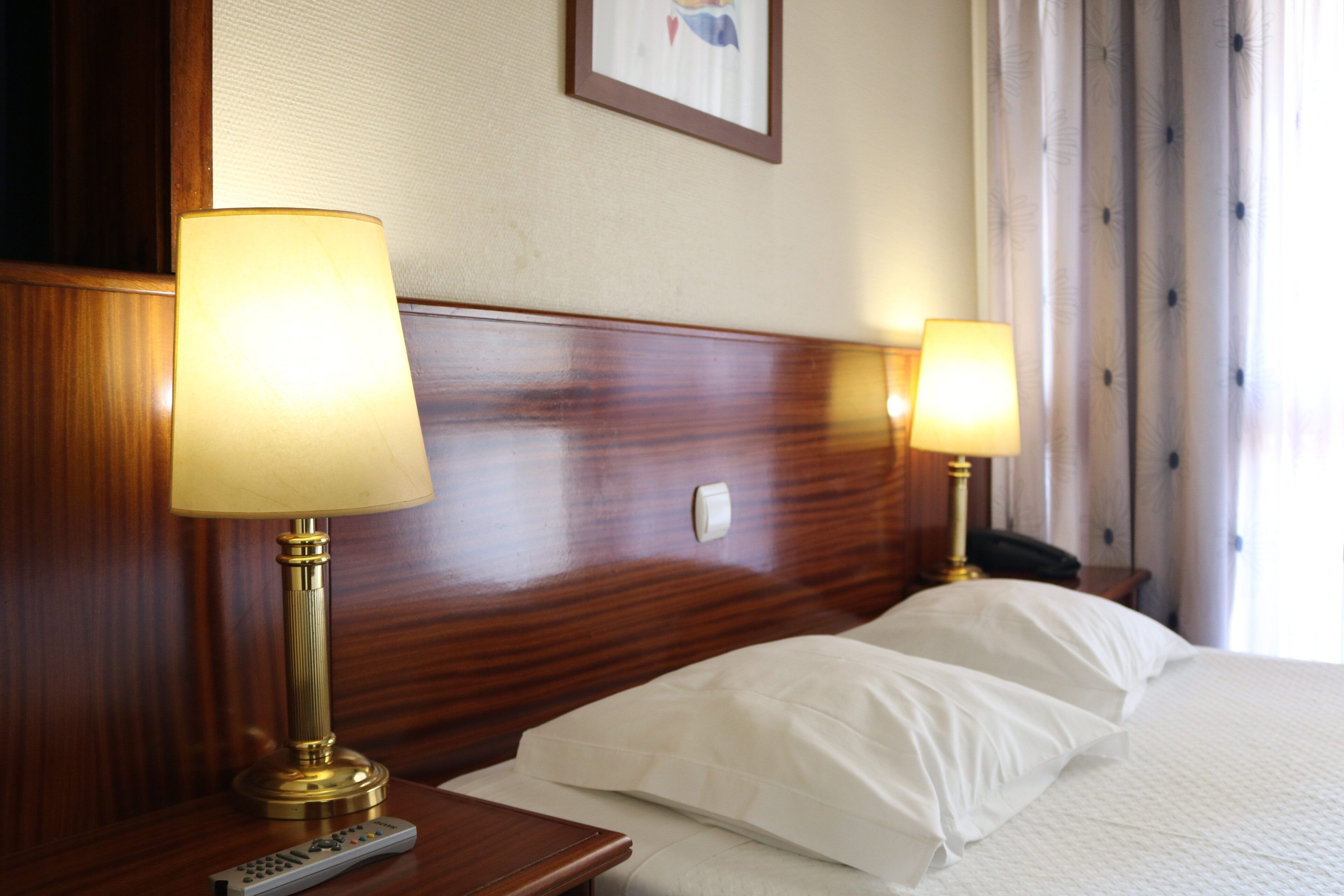 Une chambre d’hôtel avec une tête de lit en bois, deux lampes de chevet, une télécommande et un lit avec du linge de lit blanc.