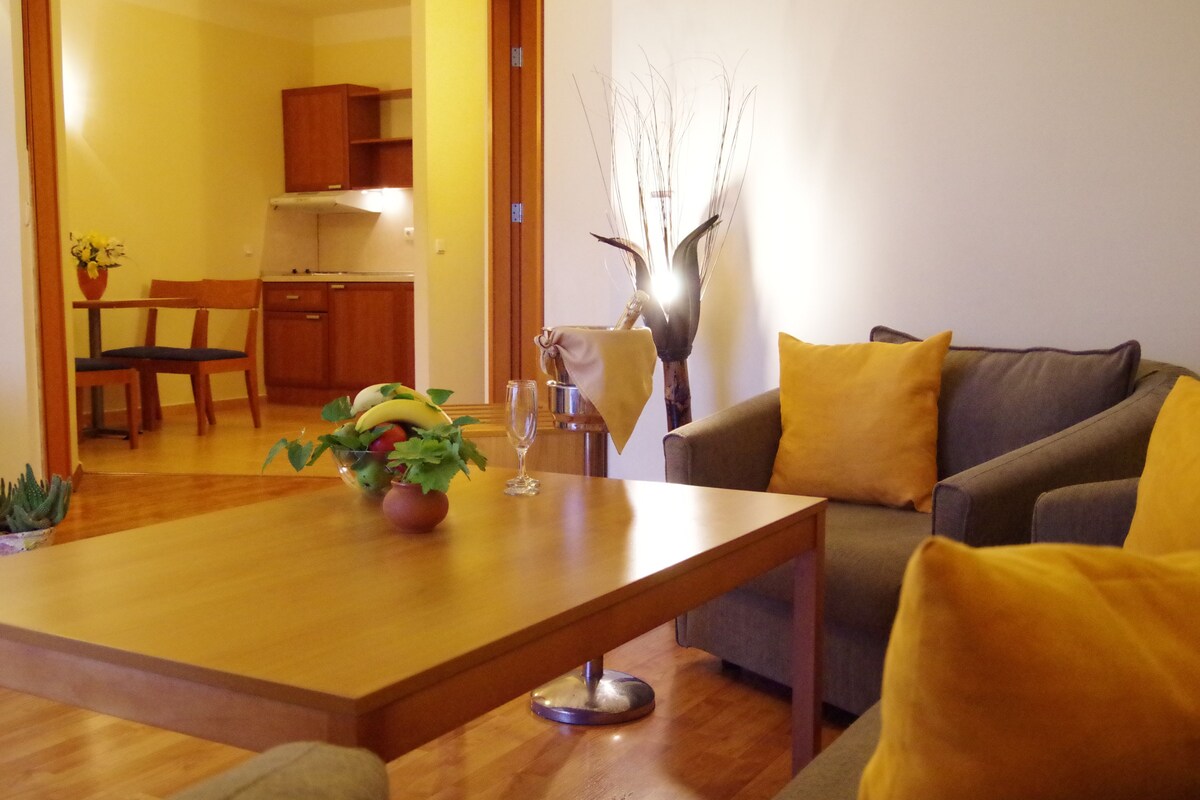 Een woonkamer met een houten salontafel, een bank met gele kussens, een vaas met takken, en een eethoek met houten meubels.
