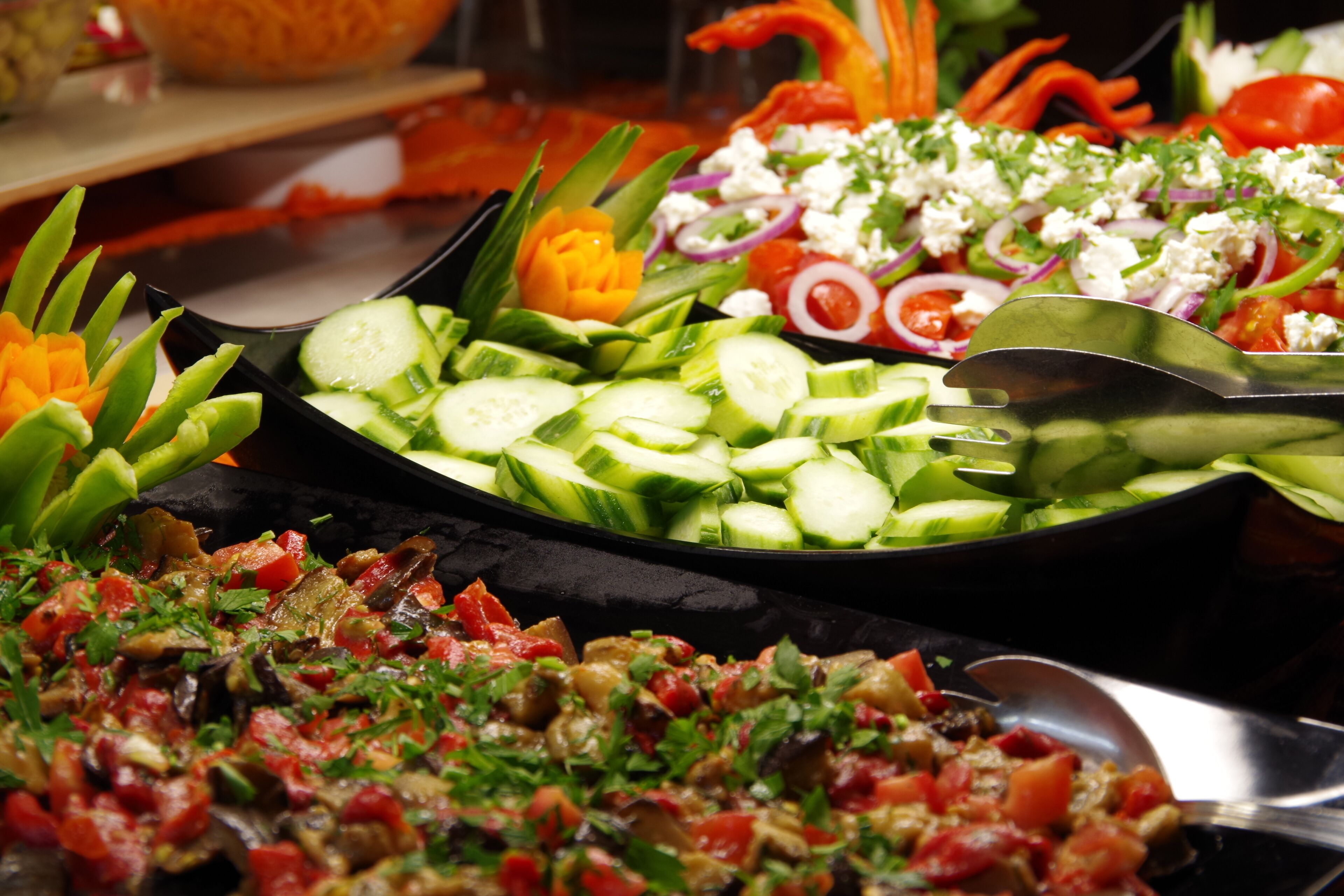 A buffet with various dishes including sliced cucumbers, a salad with tomatoes and onions, and a dish with chopped vegetables and herbs.