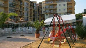 Un terrain de jeux avec des balançoires et un espace sablonneux, entouré d’un immeuble à plusieurs étages doté de balcons.
