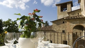 A table set outdoors with wine glasses and a white vase with flowers, set against a stone building with a bell tower.