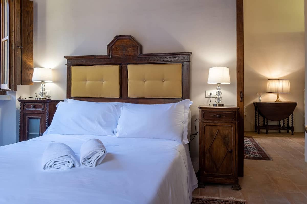 Una cama bien tendida con sábanas y toallas blancas, un cabecero de madera con dos paneles, dos mesitas de noche con lámparas y un armario de madera.