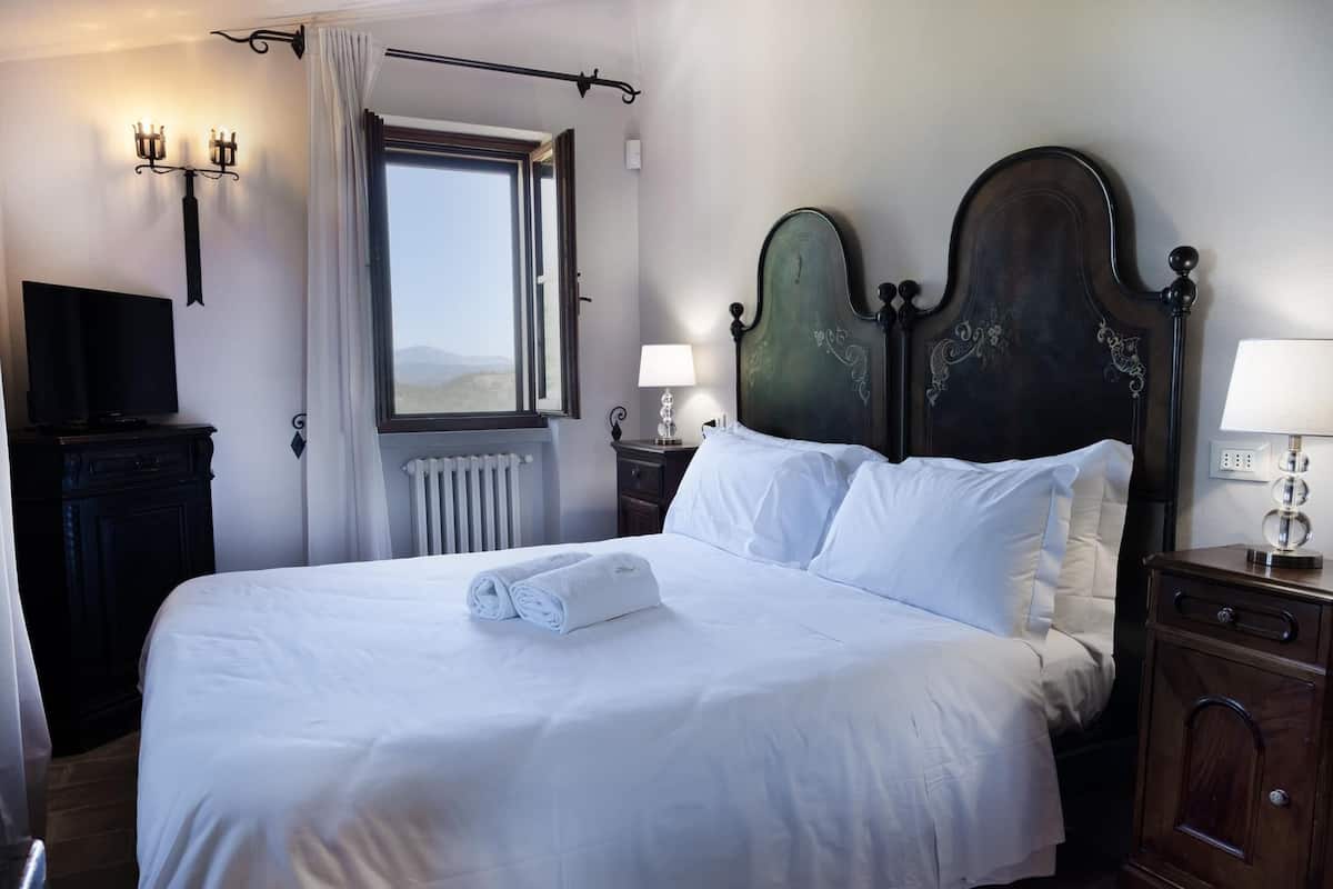 Una cama bien hecha con sábanas blancas y una toalla enrollada, un cabecero de madera oscura con detalles decorativos, una ventana con vistas al exterior y un televisor montado en la pared.