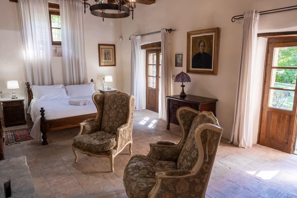 Un dormitorio con una cama, dos sillones, una mesita de noche, un aparador y una puerta que da a otra habitación.