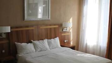 A hotel room with a large bed, wooden headboard, a bedside table with a lamp, a mirror on the wall, and a window with curtains.