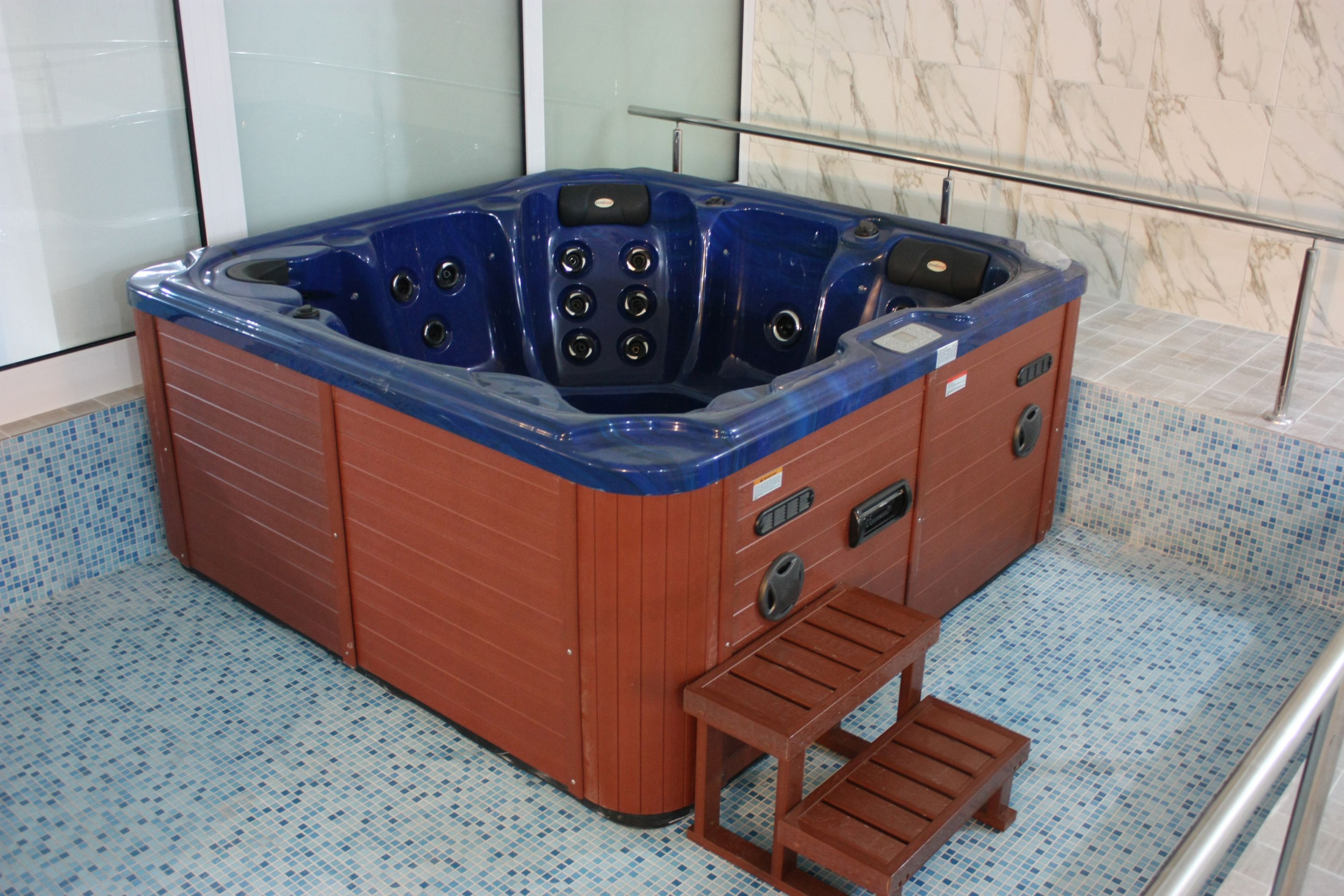 A hot tub with a wooden bench and a control panel, situated in a room with tiled flooring and a glass railing.