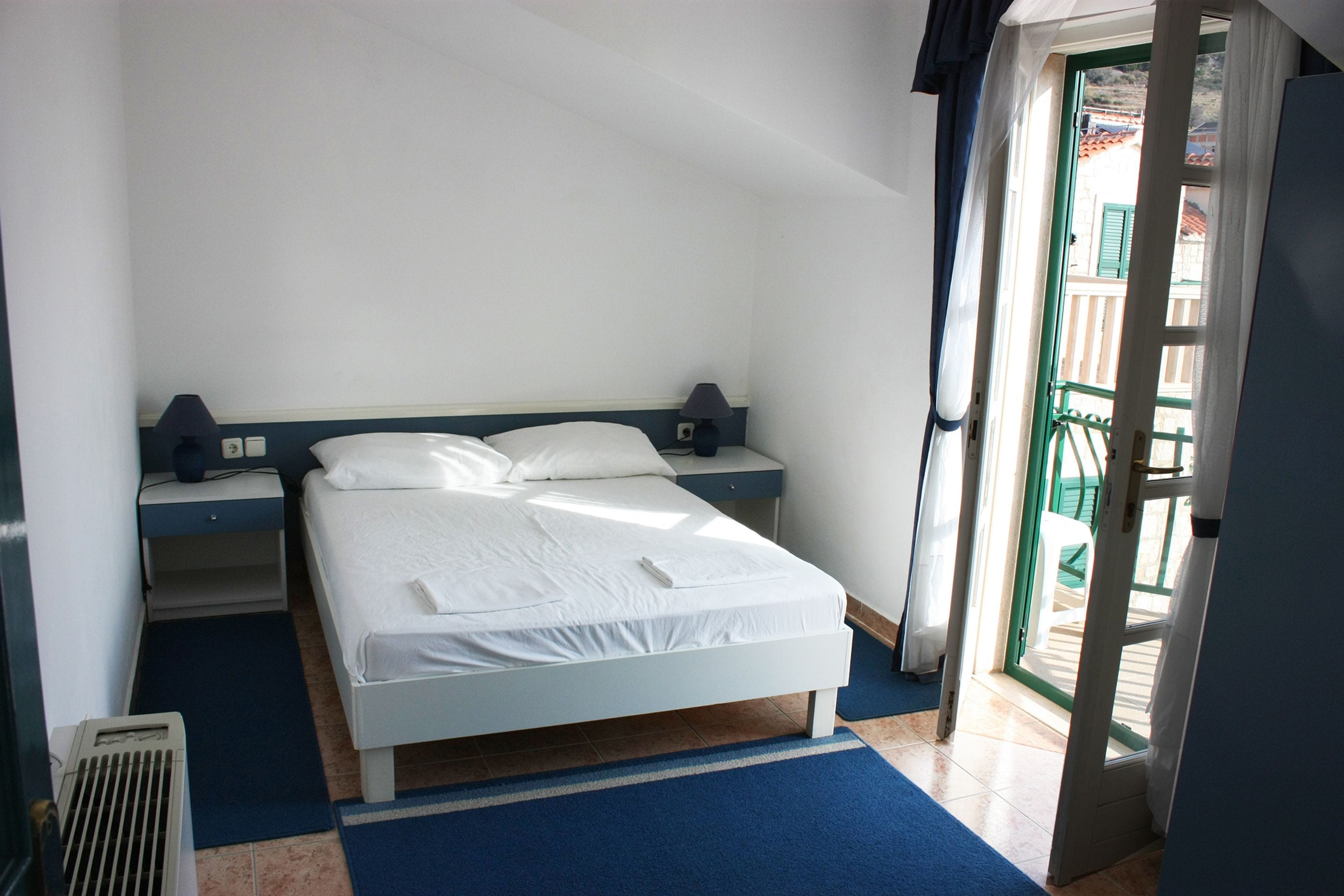A bedroom with a bed, two bedside tables, a heater, and a view of the outside.