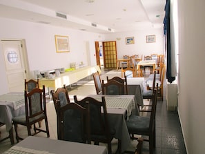 Un comedor con varias mesas y sillas, una zona de cocina con mostrador y electrodomésticos, y una puerta que da a otra habitación.