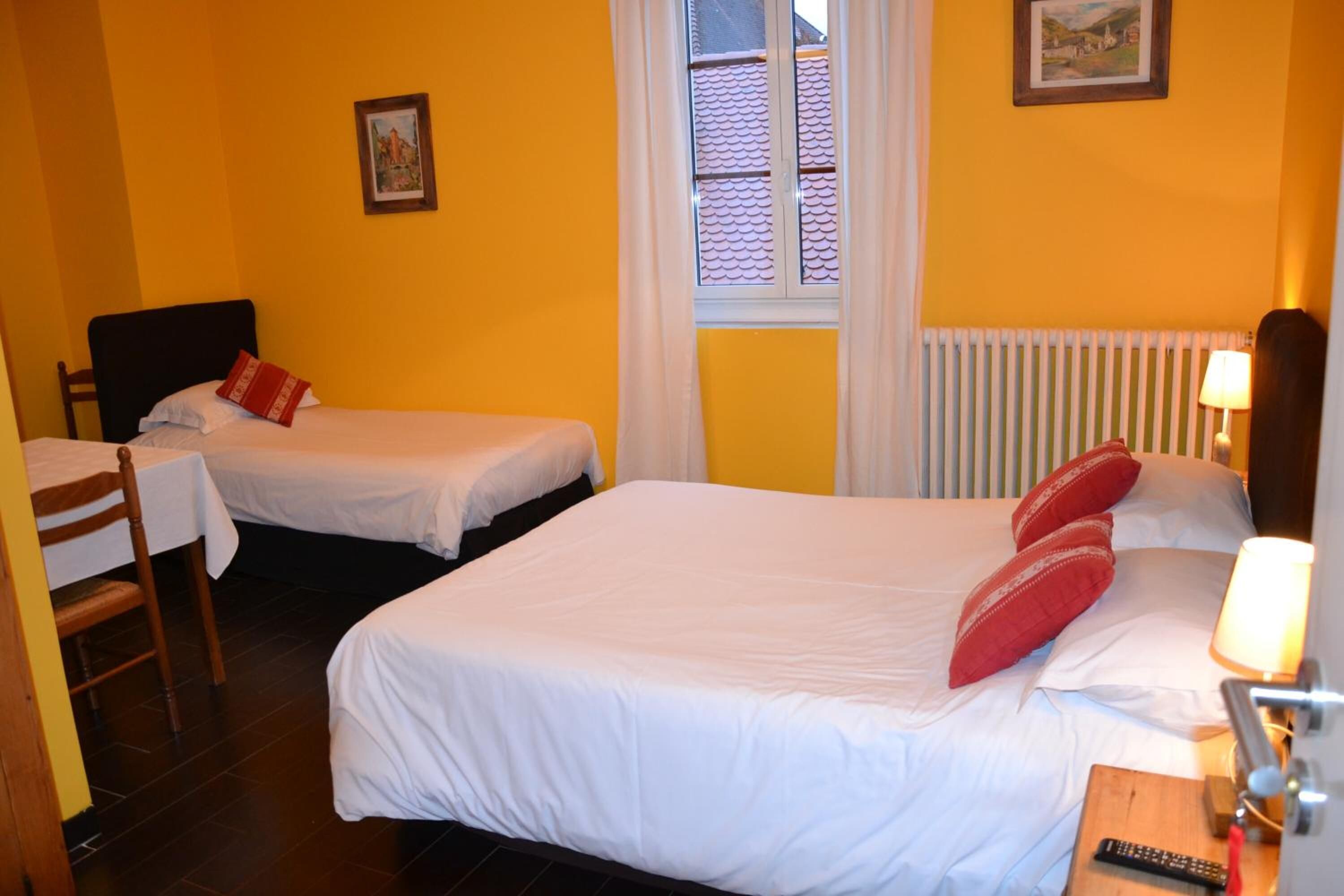 A hotel room with two single beds, a wooden chair, a small table with a lamp, and a window with a view of a building with a tiled roof.