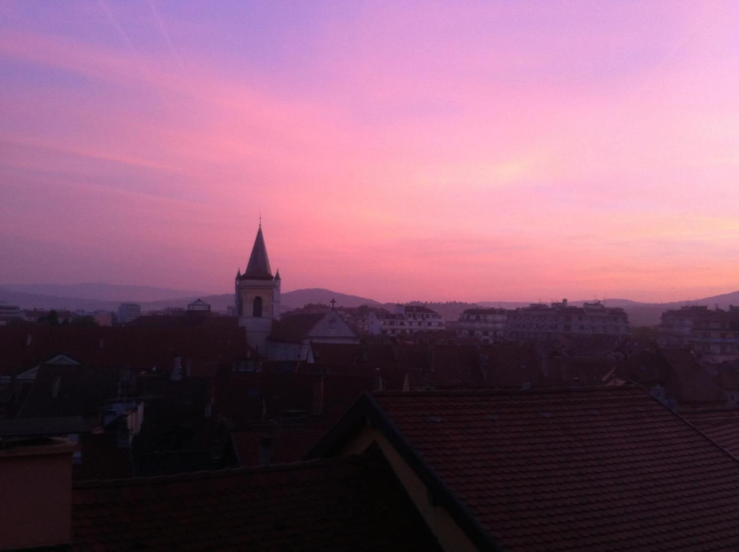 A view of a town with a prominent church tower and surrounding buildings during sunset.