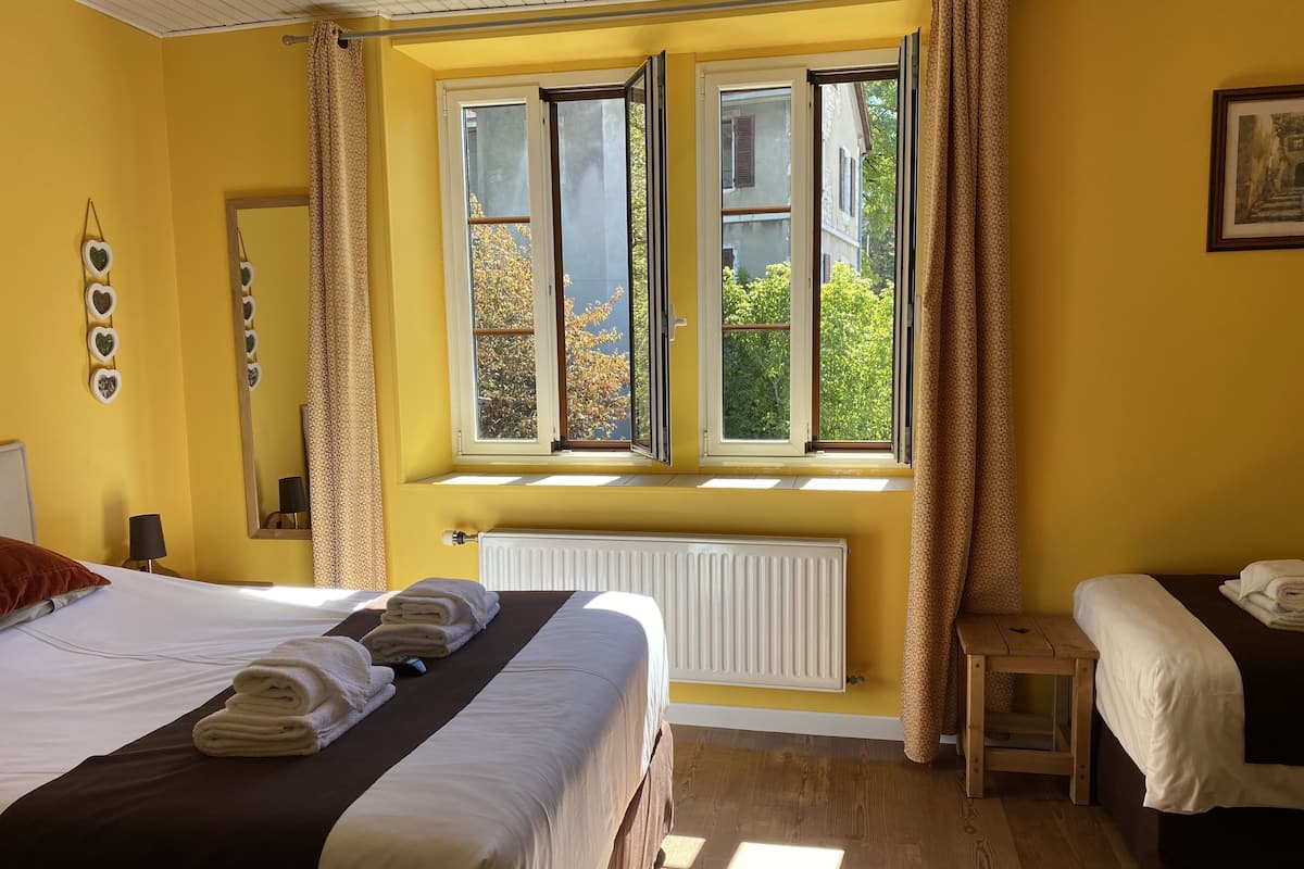Habitación de hotel con una cama grande, una cama más pequeña, una ventana con cortinas, un radiador y un espejo en la pared.