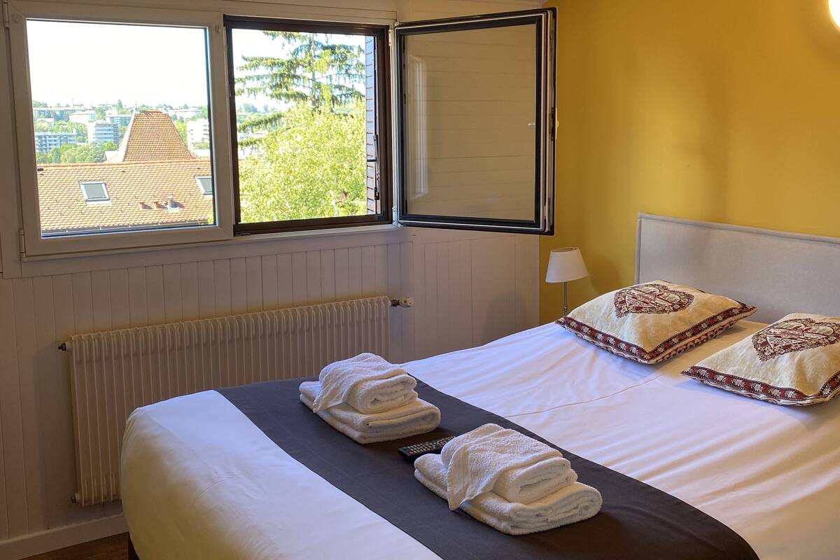 Habitación de hotel con una cama, dos ventanas, un radiador y vistas a edificios y árboles.