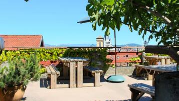 A rooftop terrace with wooden tables and benches, a potted plant, and a view of a building with a red roof and a tower.