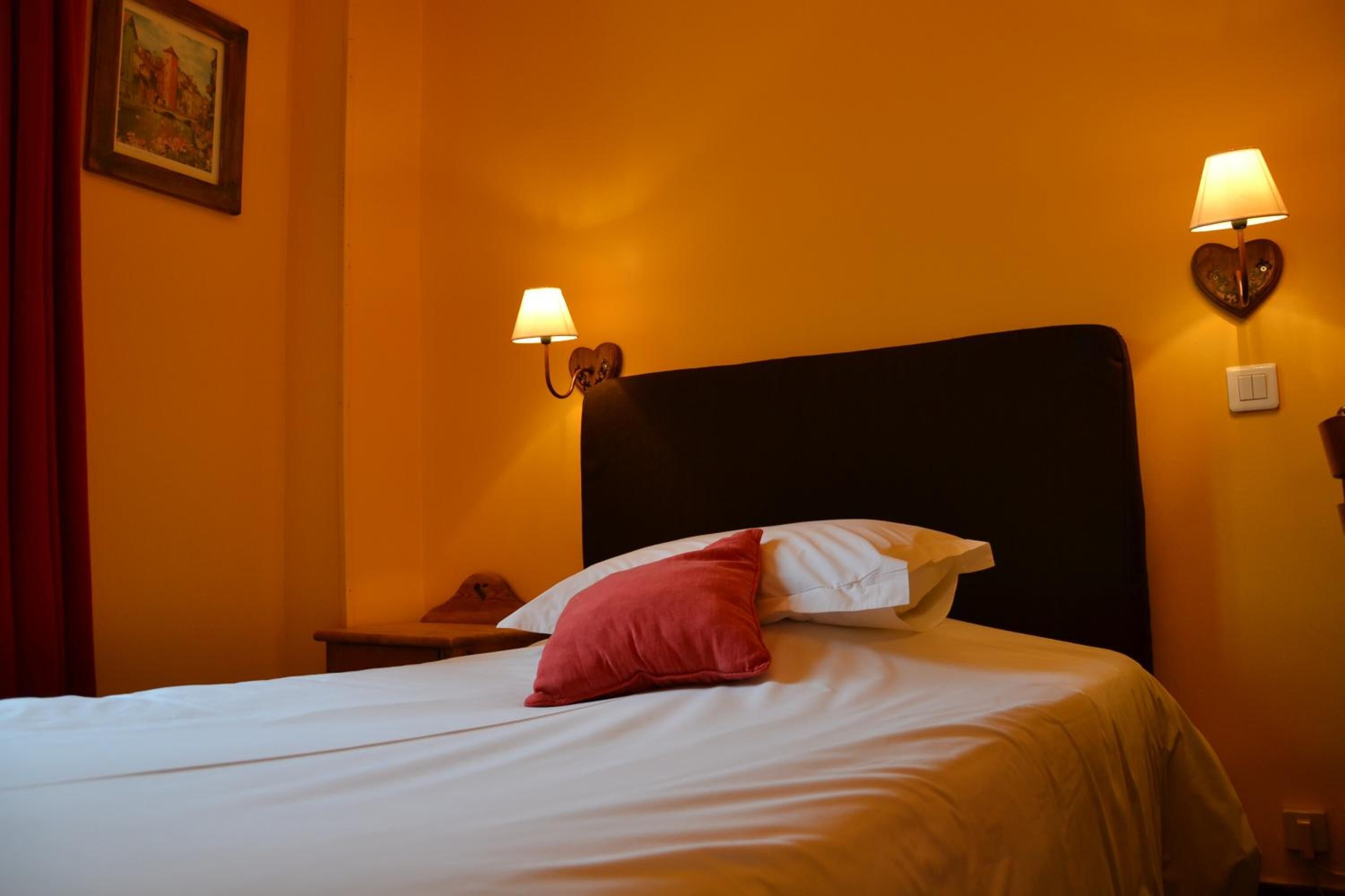 Un lit bien fait, avec des oreillers blancs et rouges, deux lampes fixées au mur et un tableau encadré accroché au mur.