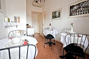 Un comedor con mesas redondas, sillas negras, manteles blancos y un estante con condimentos.