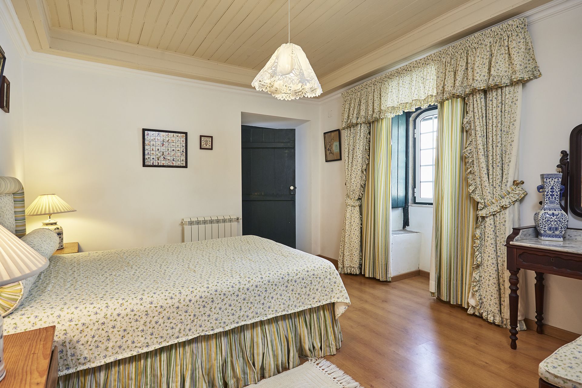 A bedroom with a bed, a bedside table, a lamp, a radiator, a door, and curtains.