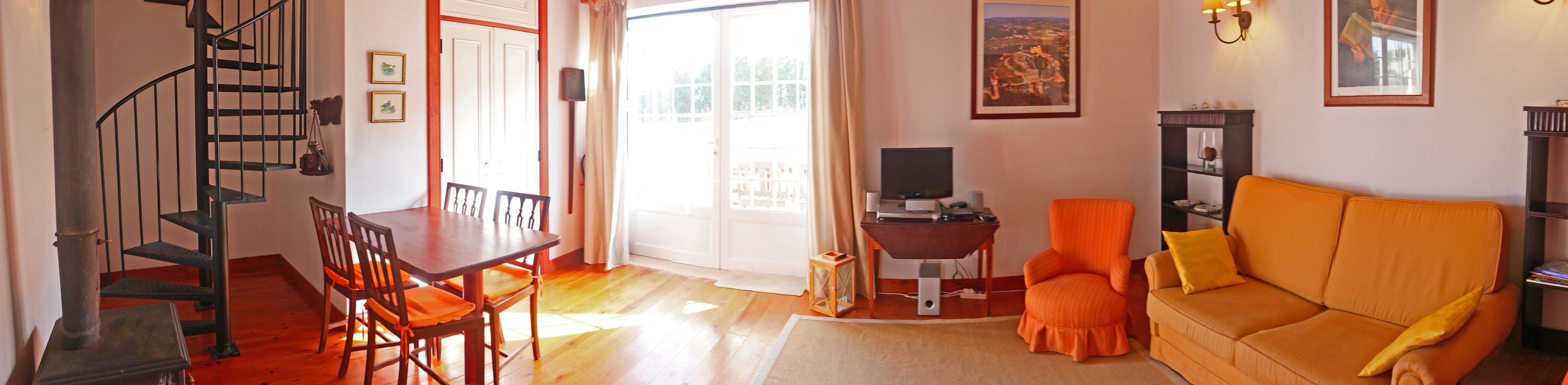 Una sala de estar con sofá, televisor, mesa de comedor con sillas, escalera y puerta que da al exterior.