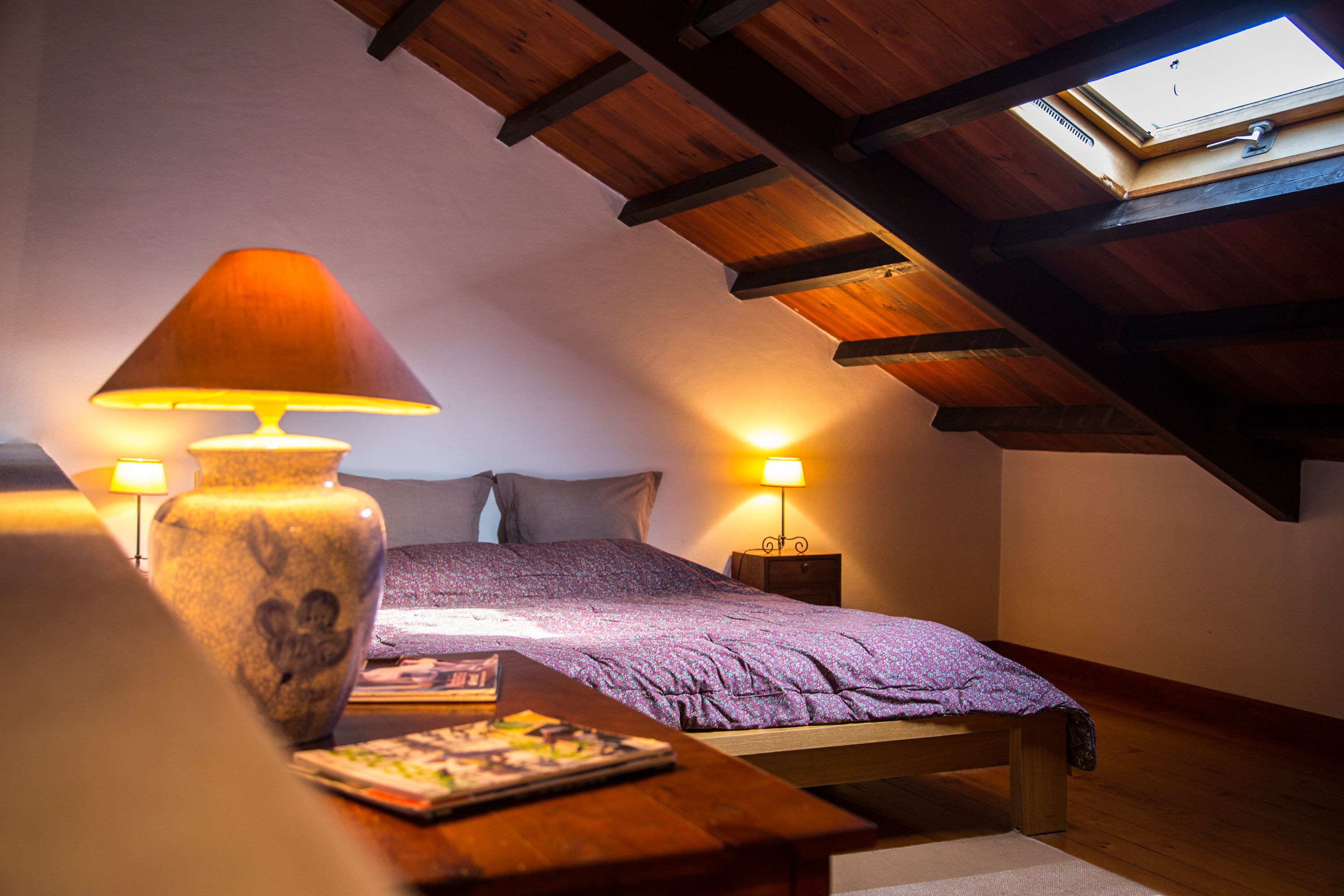 Un dormitorio acogedor con techo inclinado, una cama con manta morada, dos lámparas de noche y una mesa de madera con un jarrón y un libro.