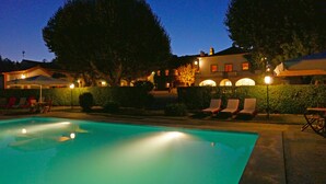 A swimming pool with lounge chairs and umbrellas, surrounded by a hedge and buildings with illuminated windows. - Quinta da Alcaidaria Mór (Ourem)
