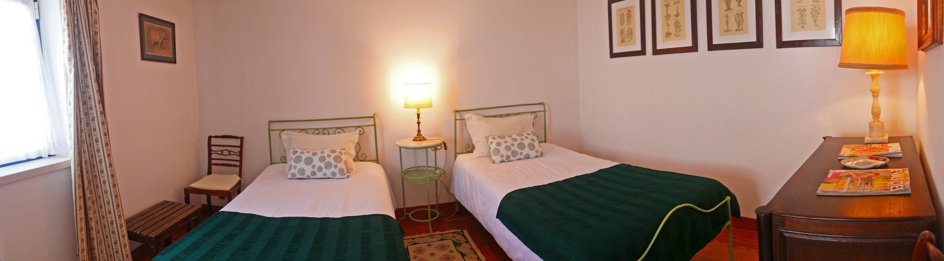 A twin room with two single beds, a small table, a chair, a lamp, and a window with curtains.