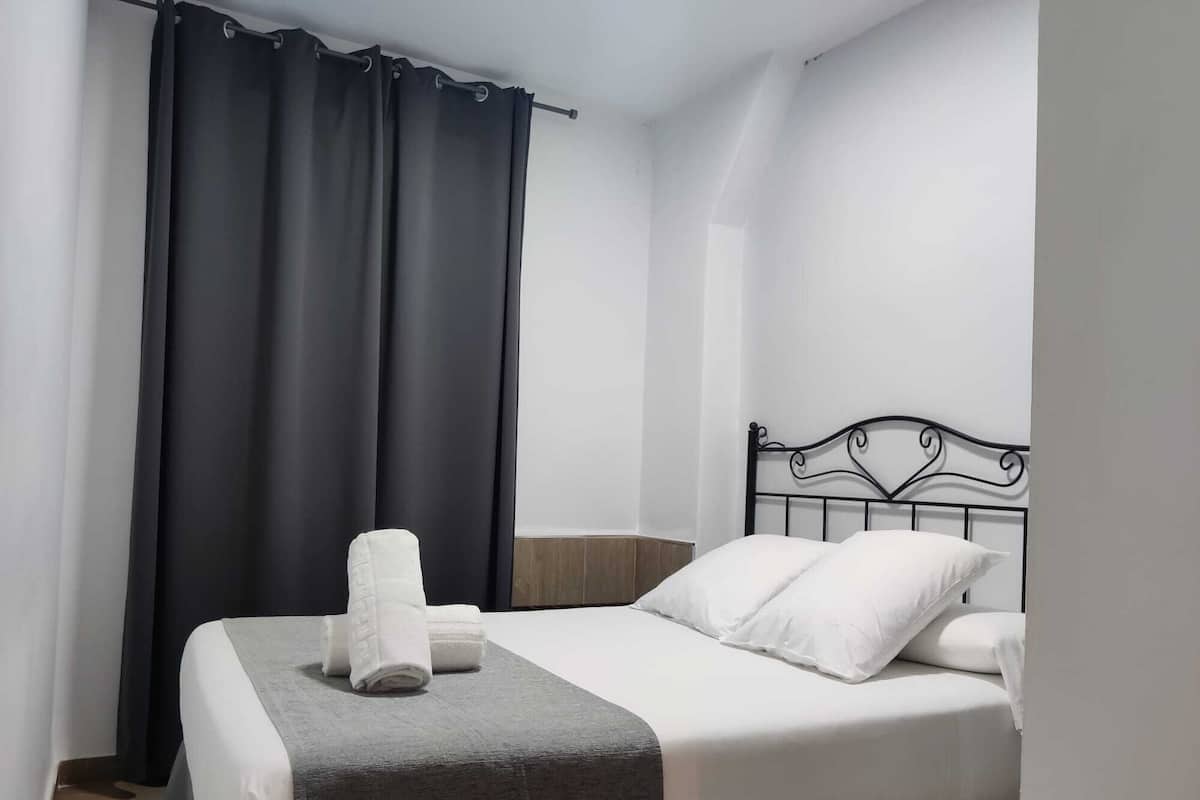 Una cama bien hecha con ropa blanca y una manta gris, cabecera de metal y una cortina oscura parcialmente abierta.