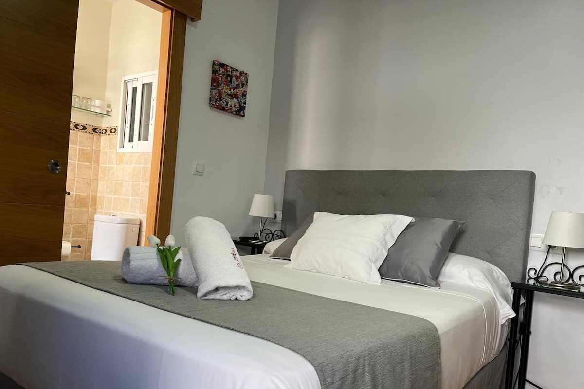 Una cama bien hecha con ropa de cama blanca y gris, un cabecero gris y una mesita de noche pequeña con una lámpara.
